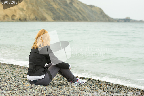 Image of A girl sits on a pebble beach by the sea in cold weather and watching the surf