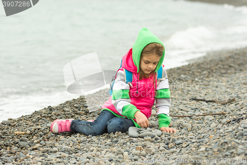 Image of Seven-year girl sitting on a pebble beach in the warm clothing and with enthusiasm plays with stones