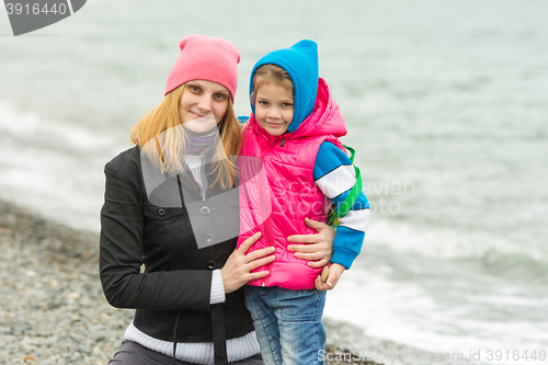 Image of Mom and daughter in warm clothes hugging each other on the beach in cold weather and with a smile look in the frame