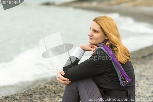 Image of A girl sits on a pebble beach by the sea on a cloudy day in cold weather, and smiles enigmatically