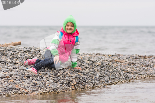 Image of Warmly dressed smiling girl sitting on the pebble near the sea and looking at the frame