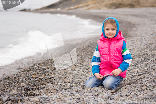 Image of Five-year girl sits on a pebble beach in the warm bright clothes on a cloudy day with a smile looks in the frame