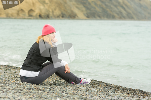 Image of A girl sits on the beach on a cloudy day in cold weather and looking thoughtfully into the distance