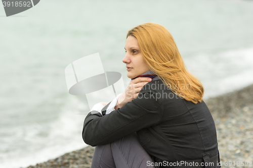 Image of A girl sits on a pebble beach by the sea on a cloudy day in cold weather, covering the neck scarf