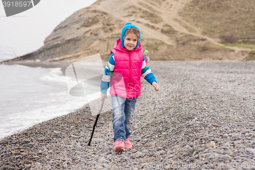 Image of Five-year girl walking on the pebble beach in the warm bright clothes with a stick in his hand