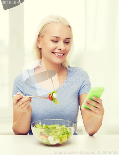 Image of smiling woman with smartphone eating salad at home