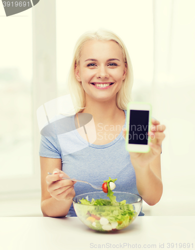 Image of smiling woman with smartphone eating salad at home