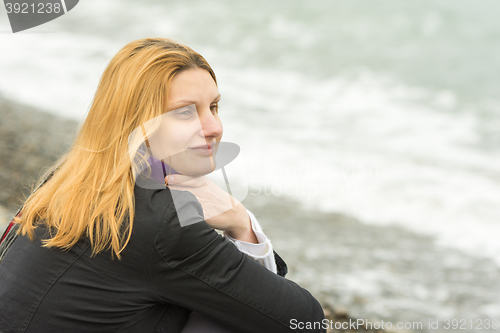 Image of A girl sits on the beach on a cloudy day in cold weather and looking thoughtfully into the distance