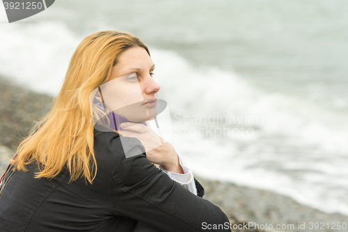 Image of A girl sits on the beach on a cloudy day in cold weather and looking thoughtfully into the distance