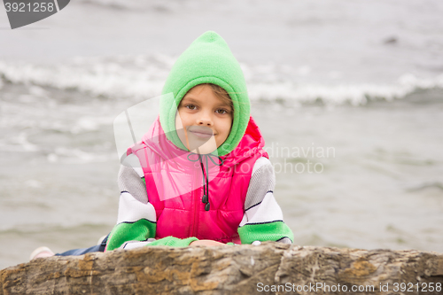 Image of Warmly dressed girl looks out from behind a rock against the backdrop of the sea on a cold cloudy day