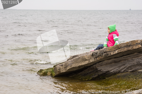 Image of Warmly dressed little girl sitting on a piece of coastal cliffs and looking at the sea