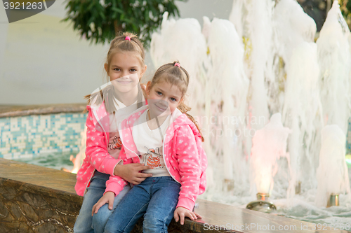 Image of Two girls sitting at the fountain in the mall and joyfully look to the Frame