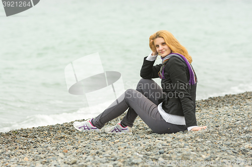 Image of  Young girl with a smile sits on a pebble beach by the sea on a cloudy cold day