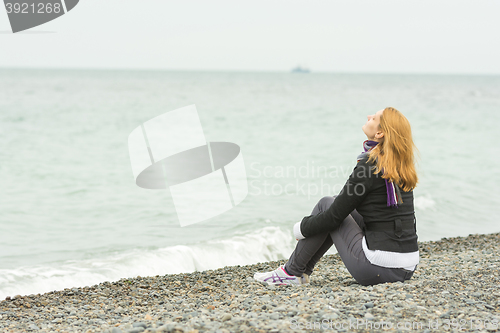 Image of Young girl sitting on a pebble beach by the sea face to the sea breeze on a cloudy day