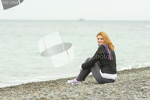 Image of  Smiling young girl sitting on a pebble beach by the sea on a cloudy cold day embracing knees