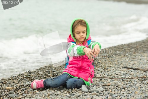 Image of Seven-year girl sitting on a pebble beach in the warm clothing and pours out through her fingers small stones