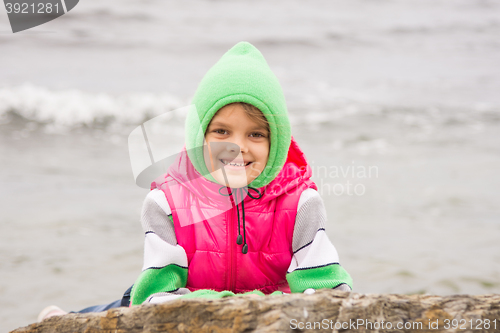 Image of Warmly dressed girl with a smile peeking out from behind a rock against the backdrop of the sea