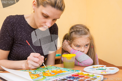 Image of The girl-artist draws on the colors of paper next to a little girl with a bored look