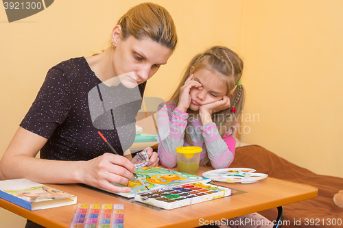 Image of Artist teacher shows how to paint watercolors, and the child rubs his eyes wearily