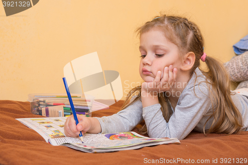 Image of Little girl studying a magazine with a pencil in his hand lying on his stomach and his head in his second hand