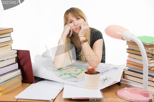 Image of The girl is sad student sitting at a table crammed with books, drawings