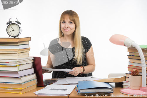 Image of The girl behind the desk littered with books with a smile, holding a paper