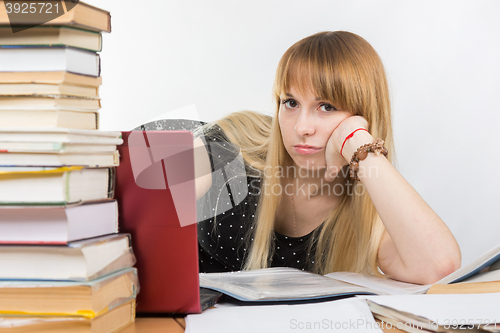 Image of Girl student sitting at a desk with a laptop and sadly looks out from behind a pile of books