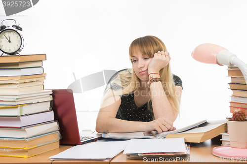 Image of Student sadly looks at the laptop at the table among the stacks of books and papers