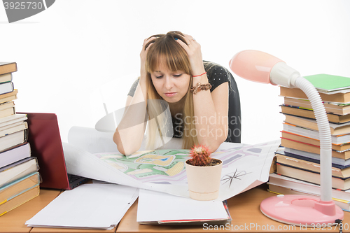 Image of A girl student at a table with a pile of books, drawings and projects sits holding his head in his hands