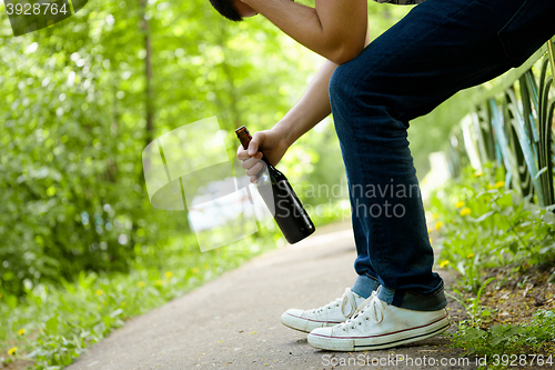 Image of Man depressed with beer bottle sitting on green fence outdoor. 