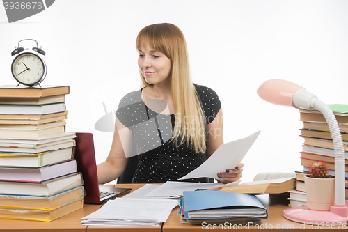 Image of Joyful girl behind a desk littered with books, papers in hand looking in laptop