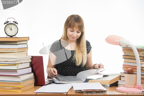 Image of A student at a table littered with books in the library with a smile, turning the pages in a folder