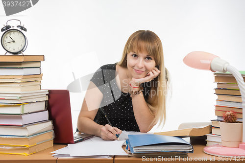 Image of  She is engaged at the table cluttered with books in the library with a smile looks in the frame