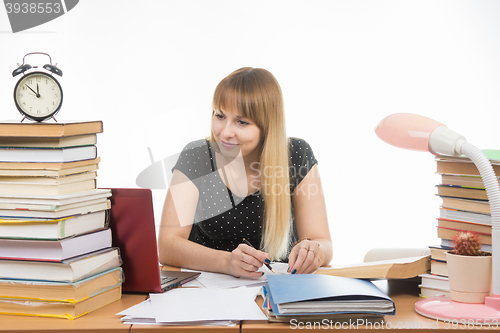 Image of A student at a table littered with books in the library with a smile looking at the laptop