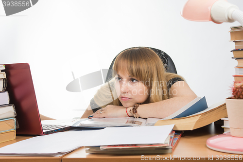 Image of The girl at the desk crammed with books and papers sadly looks at the laptop with her head resting on his hand on the table