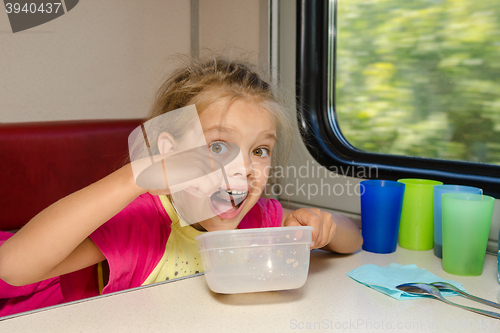 Image of The girl on the train sits at a table on the lower place in the second-class compartment of the car and eats it with relish spoon