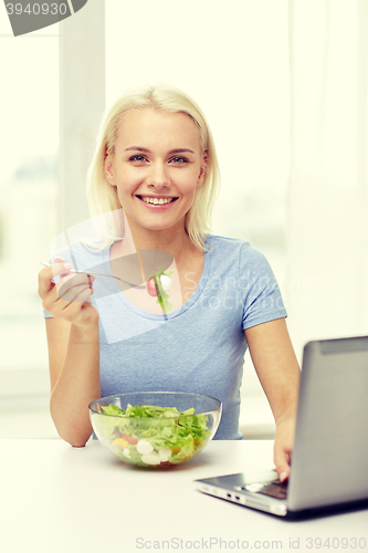 Image of smiling woman with laptop eating salad at home