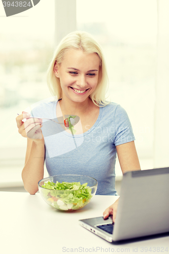 Image of smiling woman with laptop eating salad at home