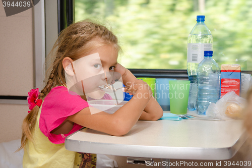 Image of A six-year girl in a train sitting at the table on the lower place in the second-class compartment of the car and eats porridge spoon