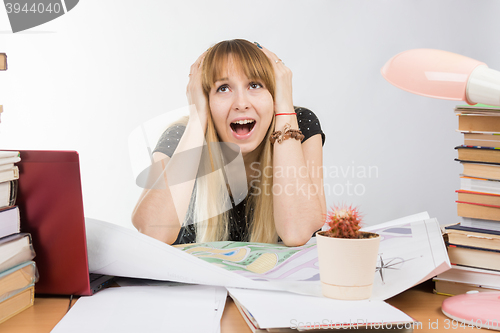 Image of Girl design student at a desk with a pile of books and projects in desperation grabbed the head