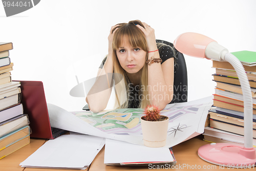 Image of Student designer behind a desk with a pile of books and projects sits holding his head in his hands