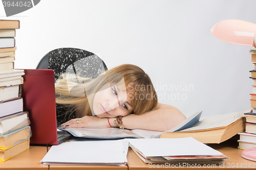 Image of Girl student laid her head on her hand crammed with books and papers lying on the desktop