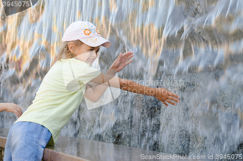 Image of Cheerful six year old girl in summer clothes hand trying to get the water artificial waterfall