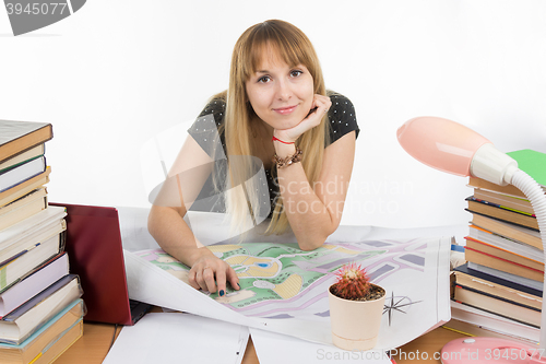 Image of A girl student at a desk littered with books and drawings with a smile looks in the frame