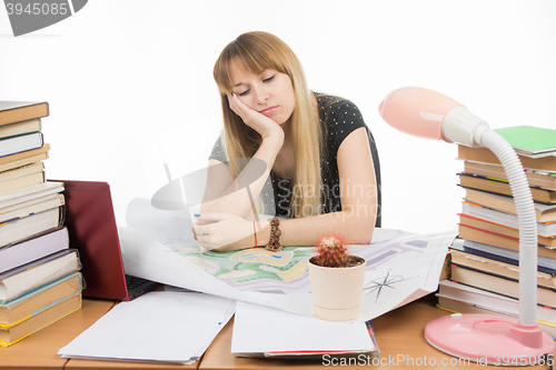 Image of A girl student at a table with a pile of books, drawings and projects sitting sadly leaning on hand and eyes closed