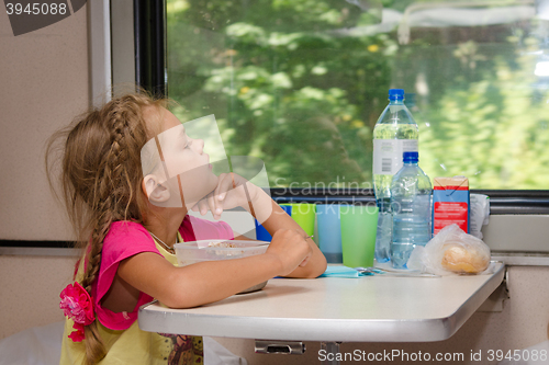 Image of A six-year girl in a train sitting at a table with food on a lower place in the second-class compartment of the car and looks thoughtfully out the window