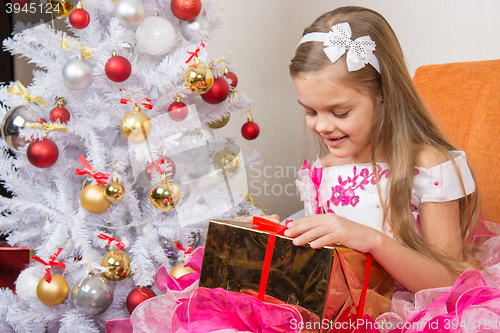 Image of Seven-year girl in a beautiful dress is considering a gift sitting on the couch at Christmas tree