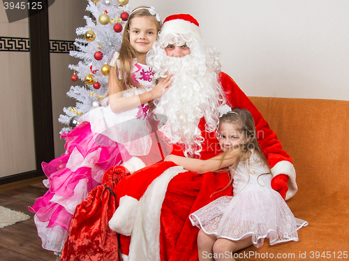 Image of Two girls in beautiful dresses hug Santa Claus sitting on a couch