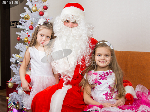 Image of Two girls in beautiful dresses hug Santa Claus sitting on the couch, one of them a little displeased