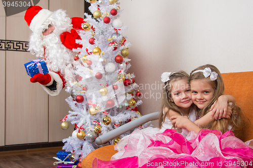 Image of Two girls hug on the couch, santa claus peeping from behind trees